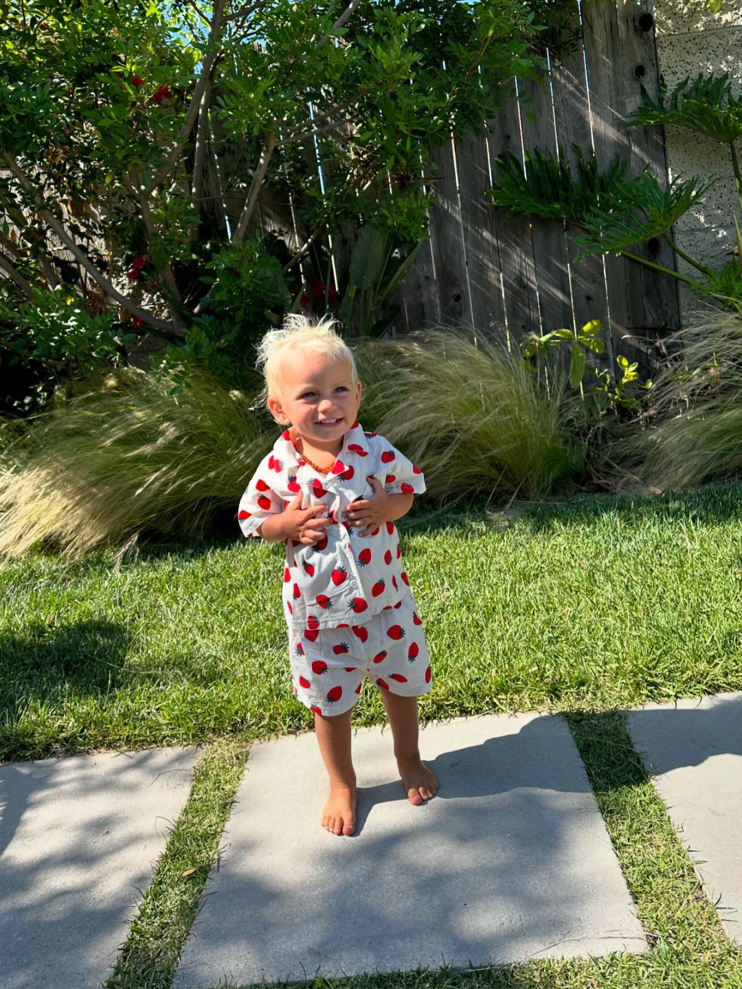 A toddler wearing the Berry Season short set in white cotton patterned with red strawberries. They are standing on a paved path in a green grassy garden in sunlight. They are smiling and have blond hair.