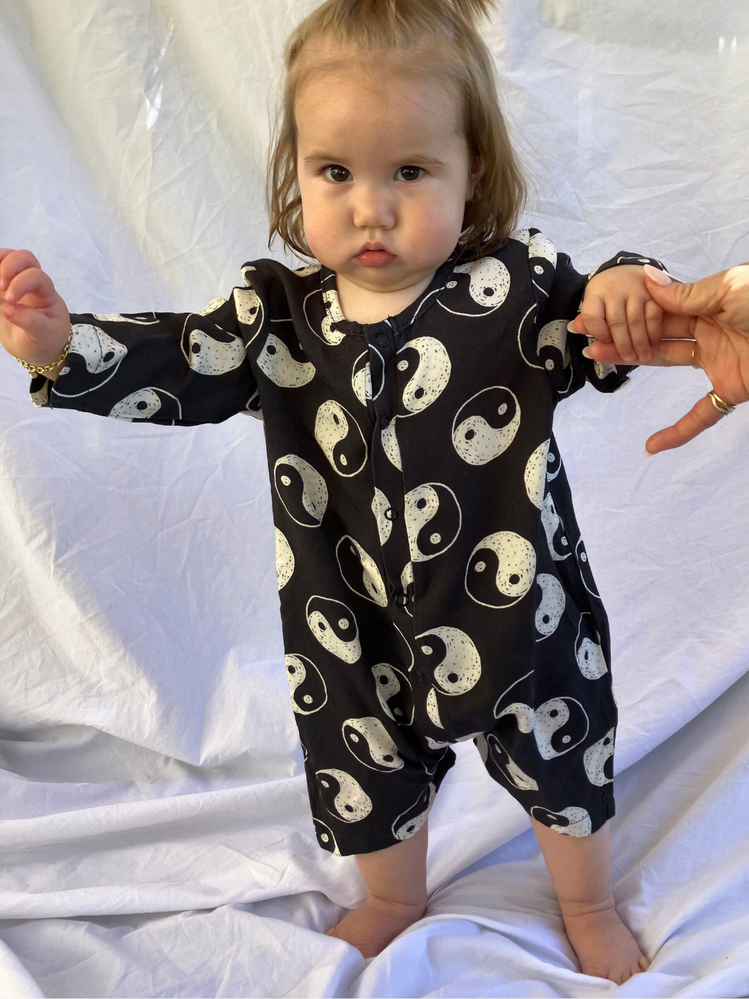 Toddler wearing the Yin Yang Baby Shortie in black cotton printed all over with white hand drawn yin yangs. She is standing holding an adult's hand, on a white sheet backdrop.