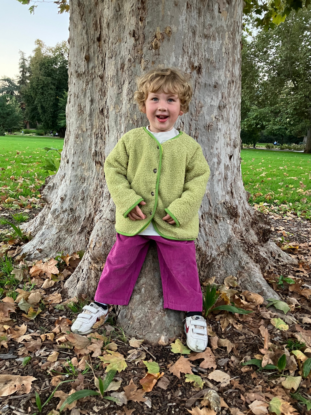 Magenta | Child in a park leaning on a tree trunk wearing magenta corduroy pants and a green fleece jacket with white velcro sneakers.