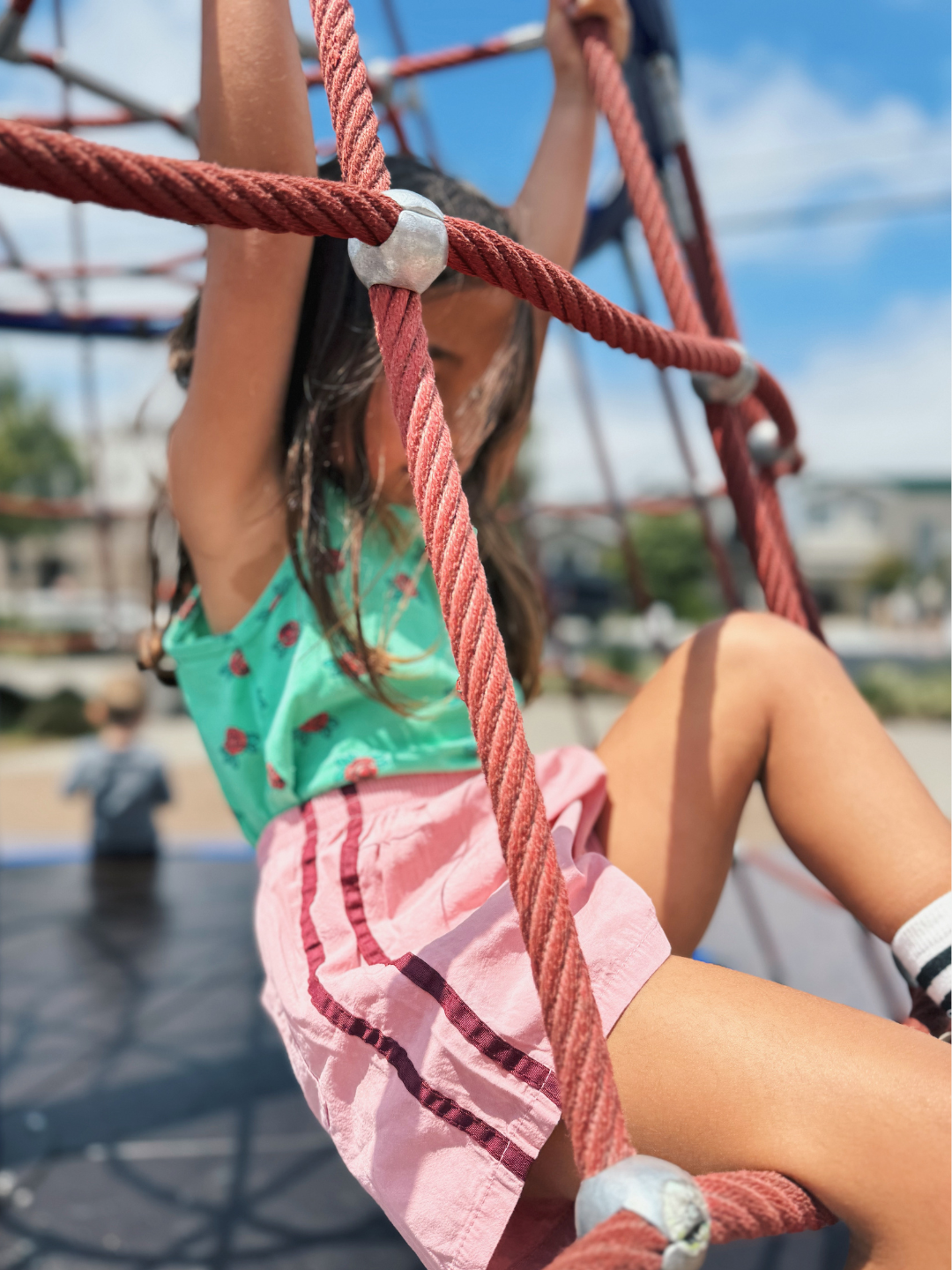Dusty Pink | Girl wearing the kids track shorts in dusty pink with maroon stripes down the side. She also wears a green tank top patterned with red roses. She is climbing on a red rope play structure.