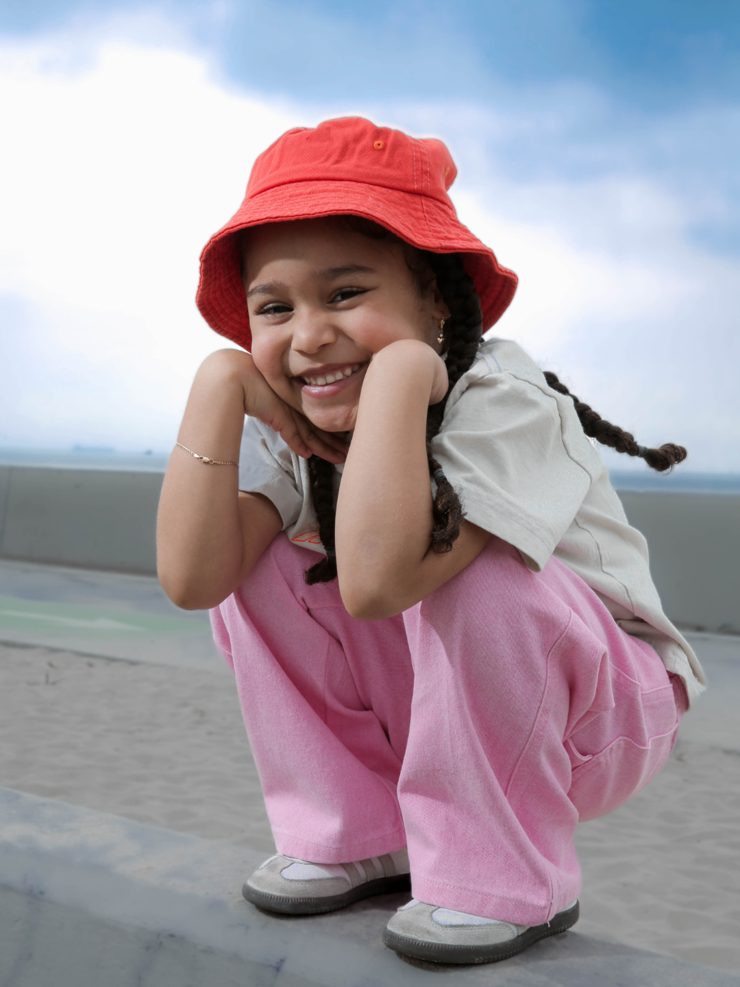 Orange | A smiling girl wearing the orange picnic bucket hat, with her hair in braids. She is squatting in pink jeans and an off-white t-shirt, and off-white sneakers, with blue sky behind her.
