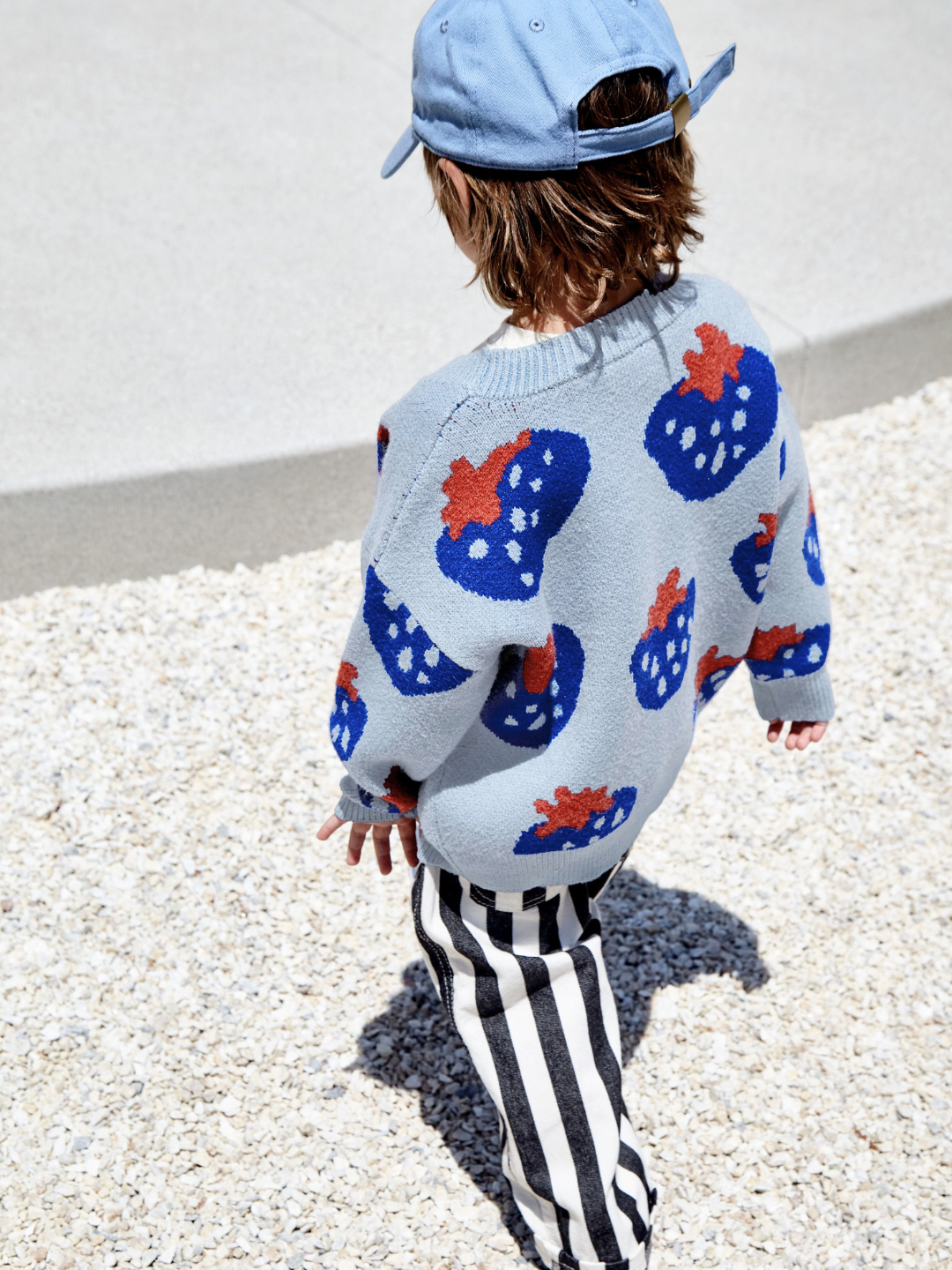 Sky | Back view of a child wearing a kids v-neck cardigan in light blue with an all-over pattern of large blue strawberries with a red leaf. He is walking on gravel in an outdoor space.