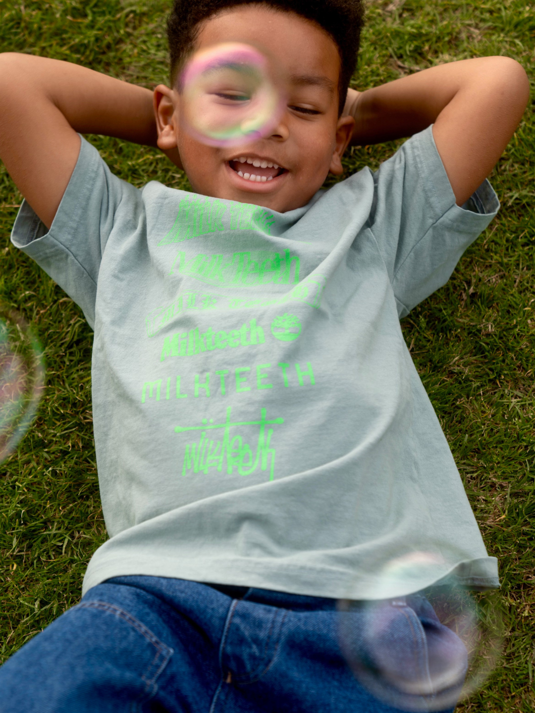 A child wearing a light green t-shirt with Milk Teeth spelled out in various fonts in neon green ink. He wears blue jeans and lies on grass, watching bubbles.