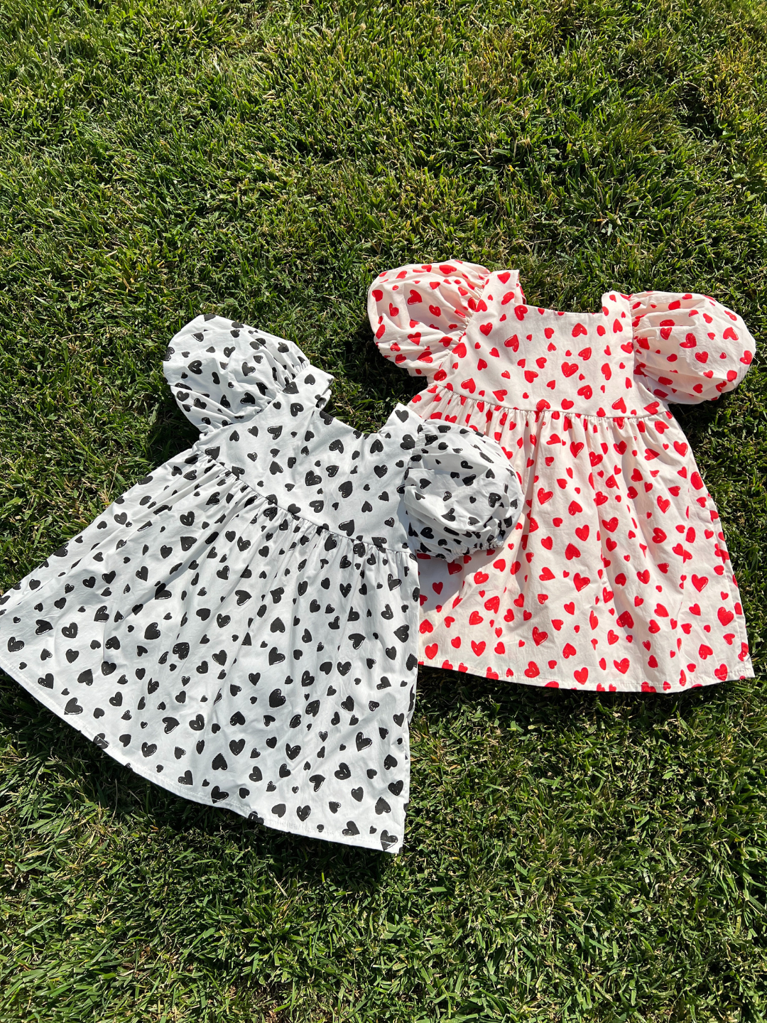 Two kids' puff-sleeved dresses laid on grass; one in a pattern of black hearts on a white background, one with red hearts