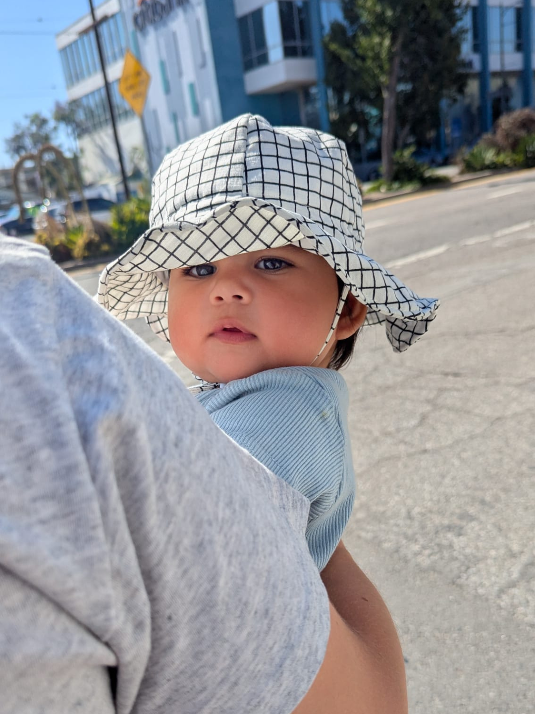 A baby wearing the Off the Grid Hat in white with a black grid check pattern. The hat has a flexible brim and a chinstrap, and the baby wears a blue top and sits in the arms of an adult in a grey tshirt, with a street scene background.