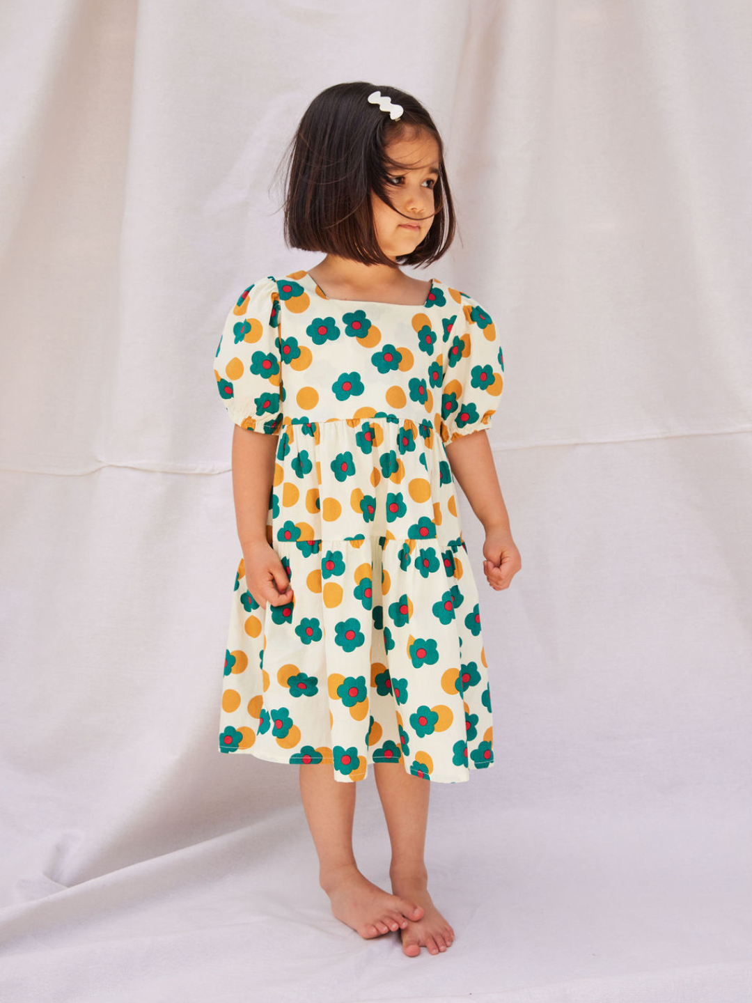 A child wearing a kids' tiered, puff-sleeved dress in a pattern of green and red flowers and ochre dots on a white background