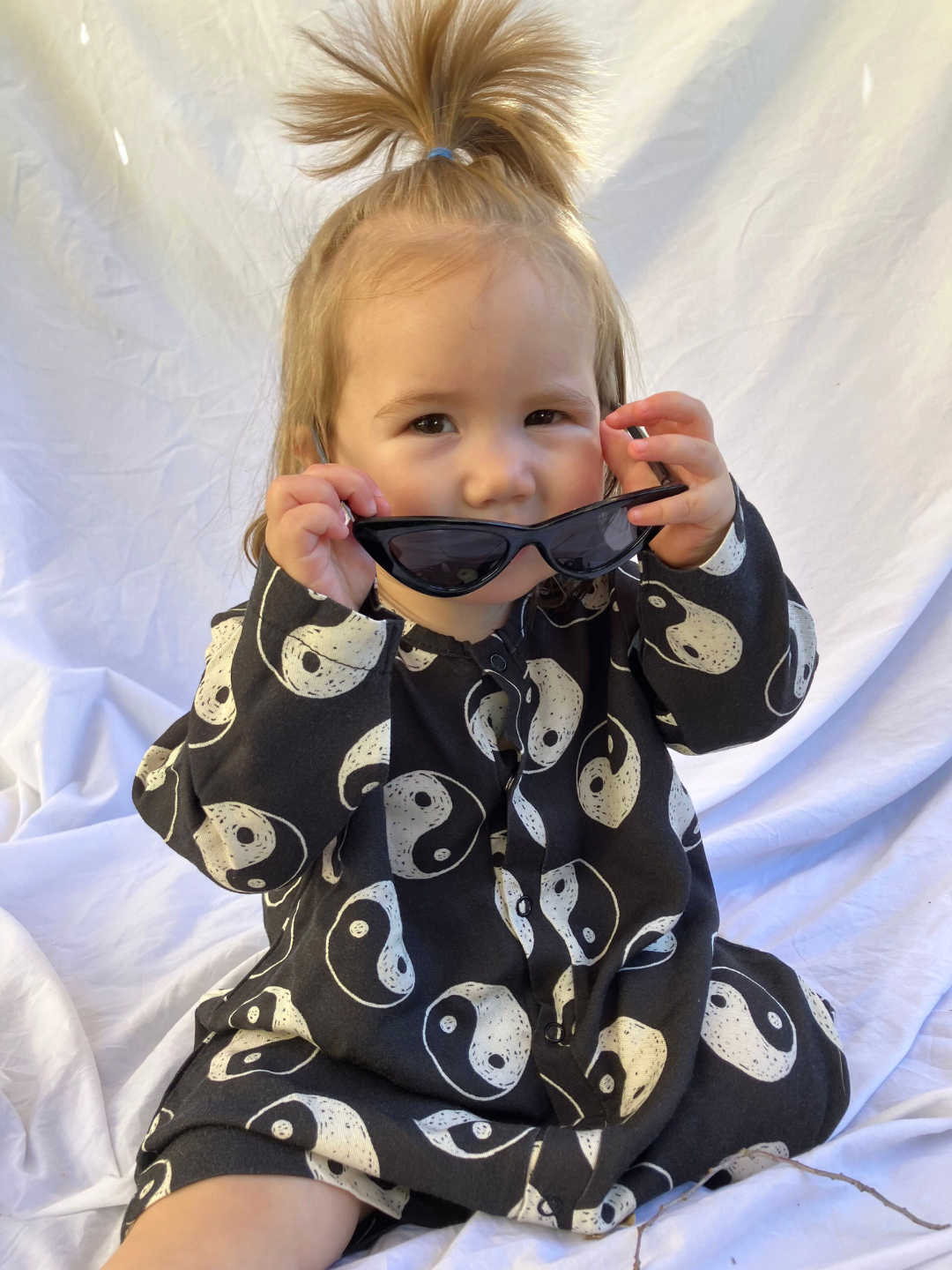 Toddler wearing the Yin Yang Baby Shortie in black cotton printed all over with white hand drawn yin yangs. She is sitting holding black sunglasses, on a white sheet backdrop and wears a top ponytail.