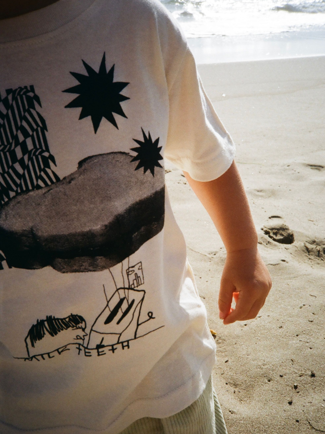 Close-up of a child on a beach wearing a kids' cream tee shirt printed with black stars, a PBJ sandwich, a toaster and the words Milk Teeth