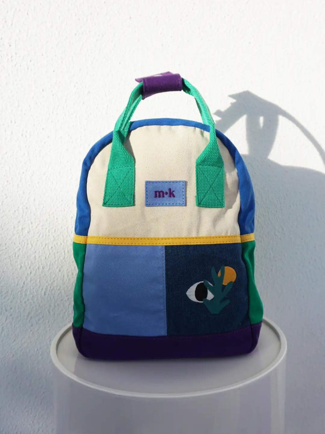 A colorblock backpack with green handles and sides, purple base, two blue patches below a cream top, one with images of an eye, a sun and a plant