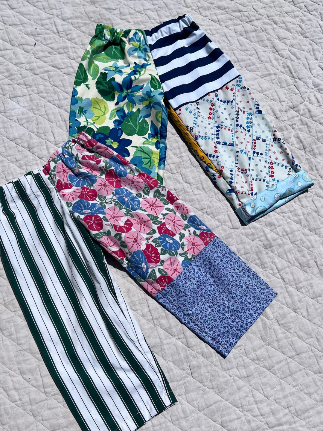Two pairs of kids' patchwork pants laid on a quilt. One pair with green and white stripes on left leg, pink and blue flowers on the right; the other with blue and green floral pattern on the left leg, stripes and diamonds on the right