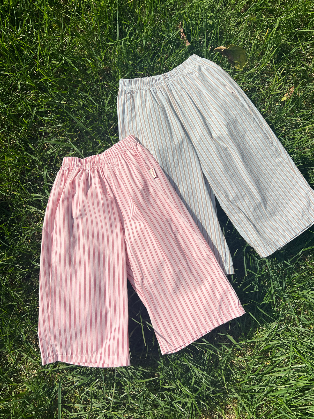 Pink | Two pairs of kids summer pants, one pink and white striped and one light blue, on grass.