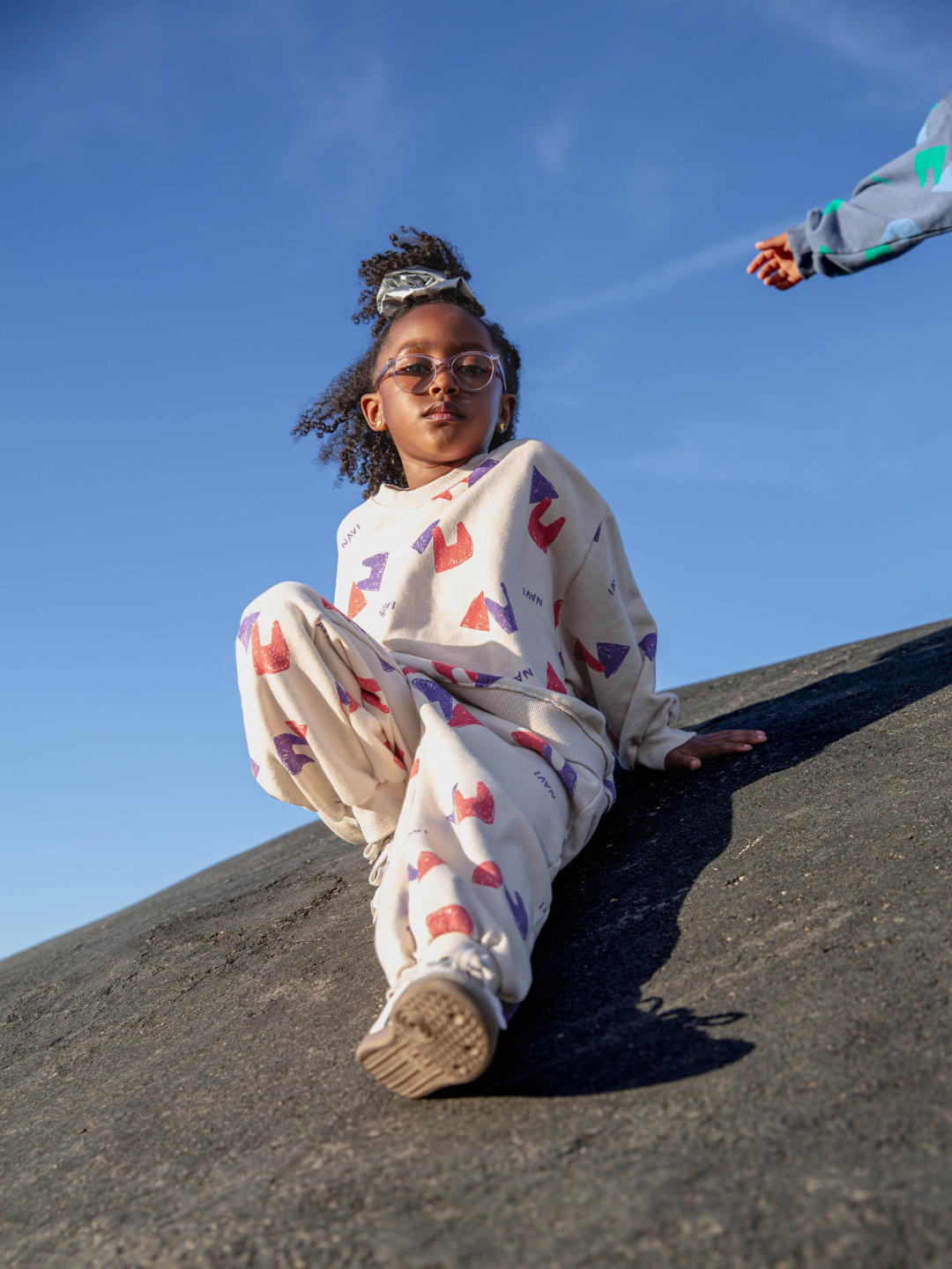 Oatmeal | Child wearing beige sweatpants with an all over pattern of red and purple shapes and the brand name Navi, with matching crewneck sweatshirt. She sits on a concrete hill with blue sky above.