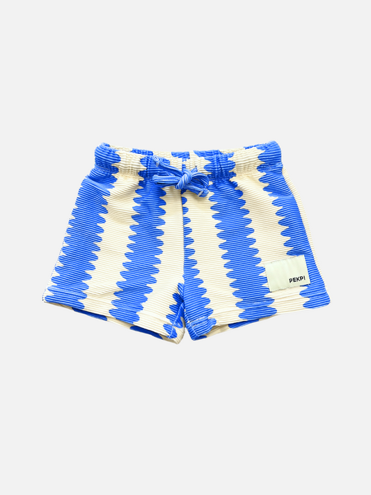 Image of SWIM SHORTS featuring a blue and white zigzag pattern, elastic waistband with front tie, and a small rectangular label on the left leg, made from recycled material.