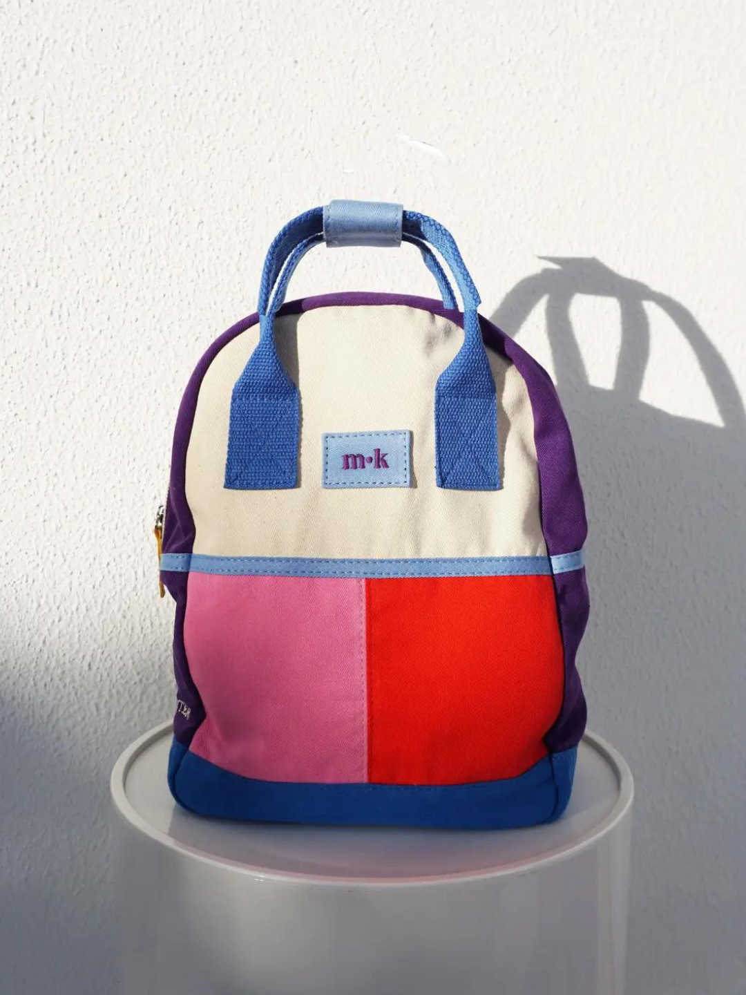 A colorblock backpack with bright blue handles and base, purple sides, and one pink, one red patch under a cream top