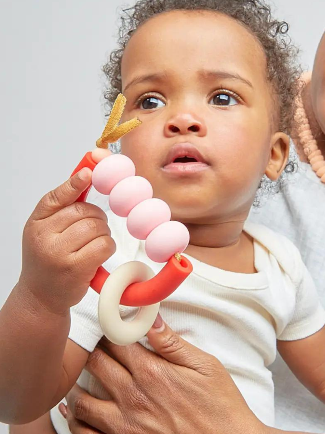 Rose | Baby holding teether with white ring on red semicircle, pale pink beads strung along top