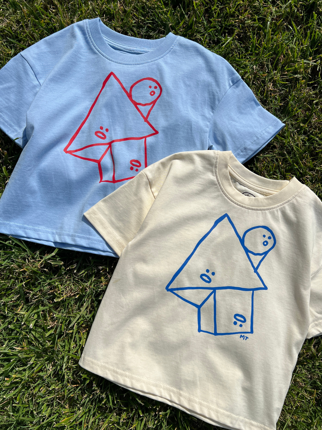 Cream | Two kids' Jumble Tees laid flat on a quilt in the sun, one cream and one sky blue