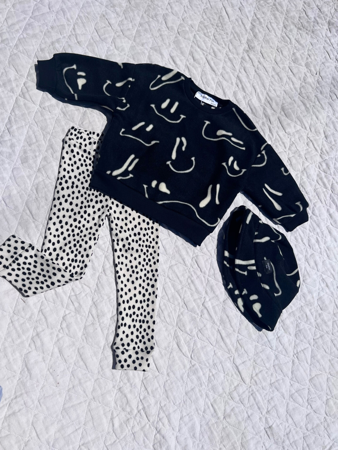 A kids' outfit laid out on a quilt, showing a pair of kids' leggings in a black and white mini cheetah dot print, with a Wavy Grin Fleece Sweatshirt and matching bucket hat in black and white