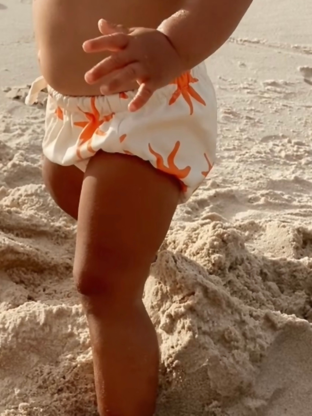 Capri | A waist-down image of a toddler wearing the Capri swim diaper in cream with orange stars all over, standing on sand.
