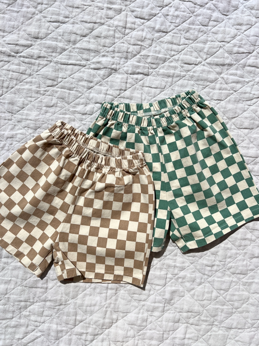 Tan | Two pars of kids Frankie shorts are laid out on a white canvas quilt. The pair on top is tan and ecru checkerboard, the pair behind is teal and ecru checkerboard.