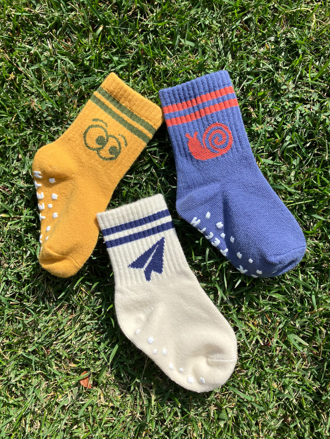 The RIBBED GRIP SOCK SET, consisting of three pairs of child essentials, is shown laid out on green grass: a yellow pair adorned with owl eyes, a blue pair featuring a snail design, and a beige pair with a paper airplane motif. These ribbed socks also come with anti-skid grips to ensure your little ones stay safe during their playful adventures.