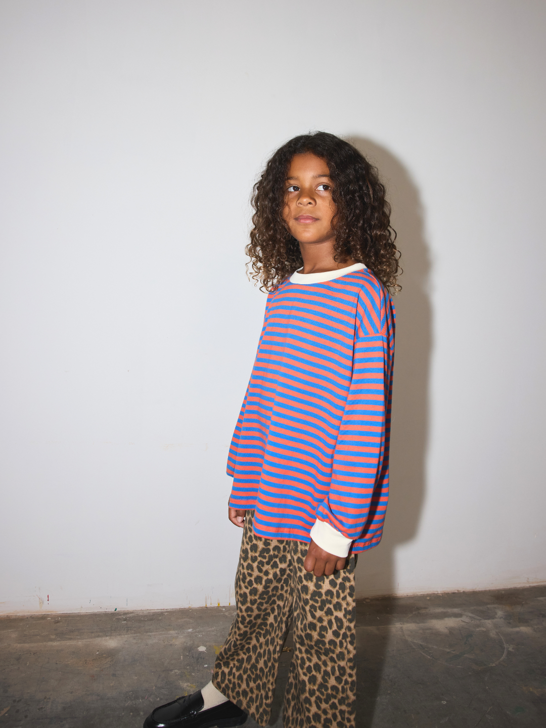 A child wearing a vibrant blue and orange striped long-sleeve top with white cuffs, paired with leopard print pants and black loafers, standing against a plain white wall.