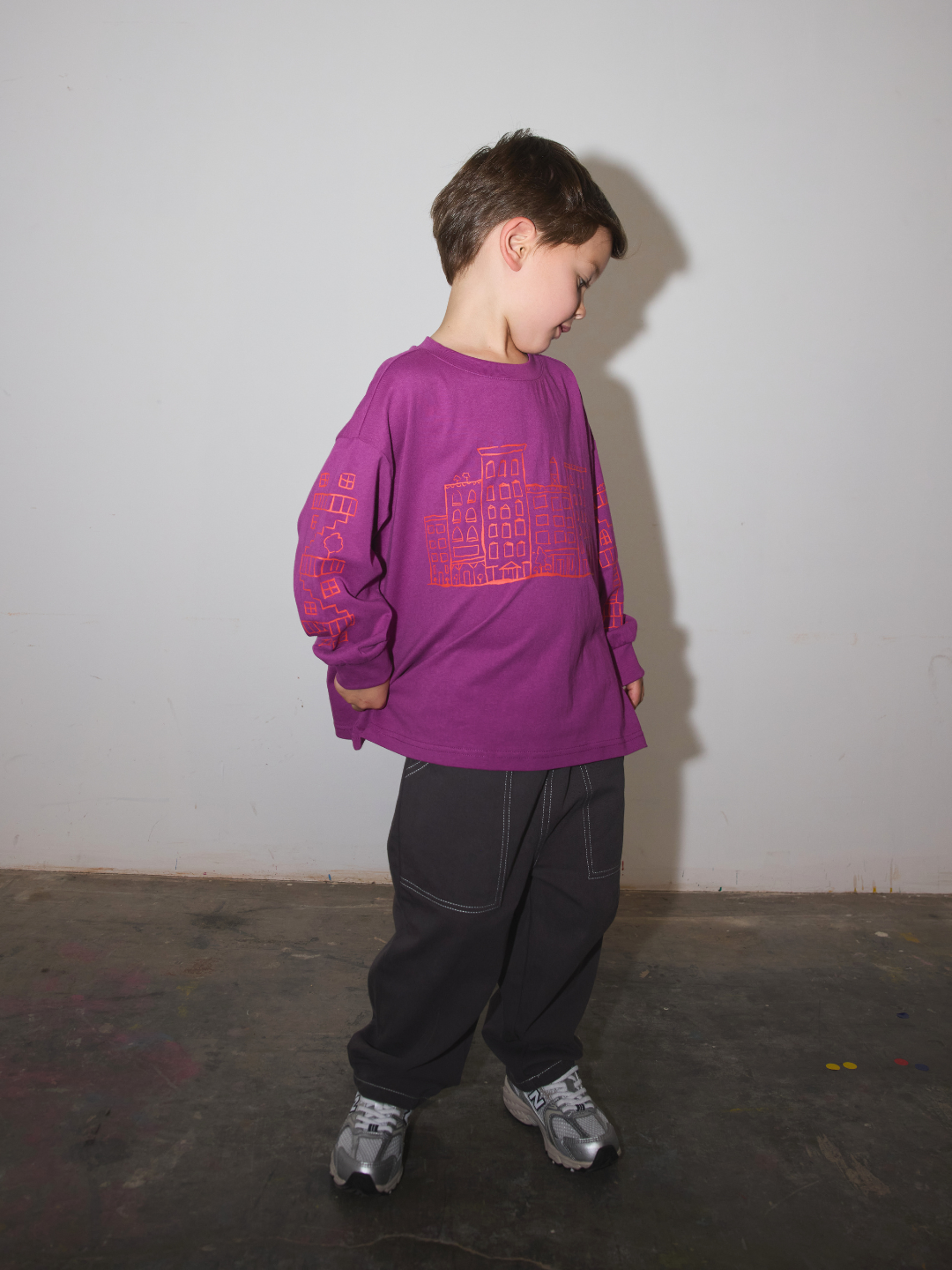 Magenta | A young boy wearing a purple long-sleeve t-shirt with a pink graphic print, black pants, and gray sneakers, standing against a plain light gray wall.