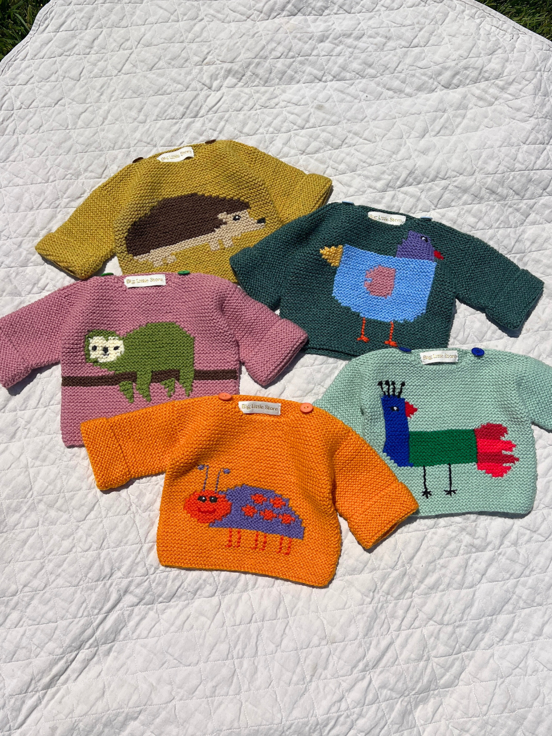 L (orange bug) | Five baby sweaters shown flat on a white quilt background: ochre with hedgehog, teal with bird, pink with sloth, mint with bird and orange with purple bug.