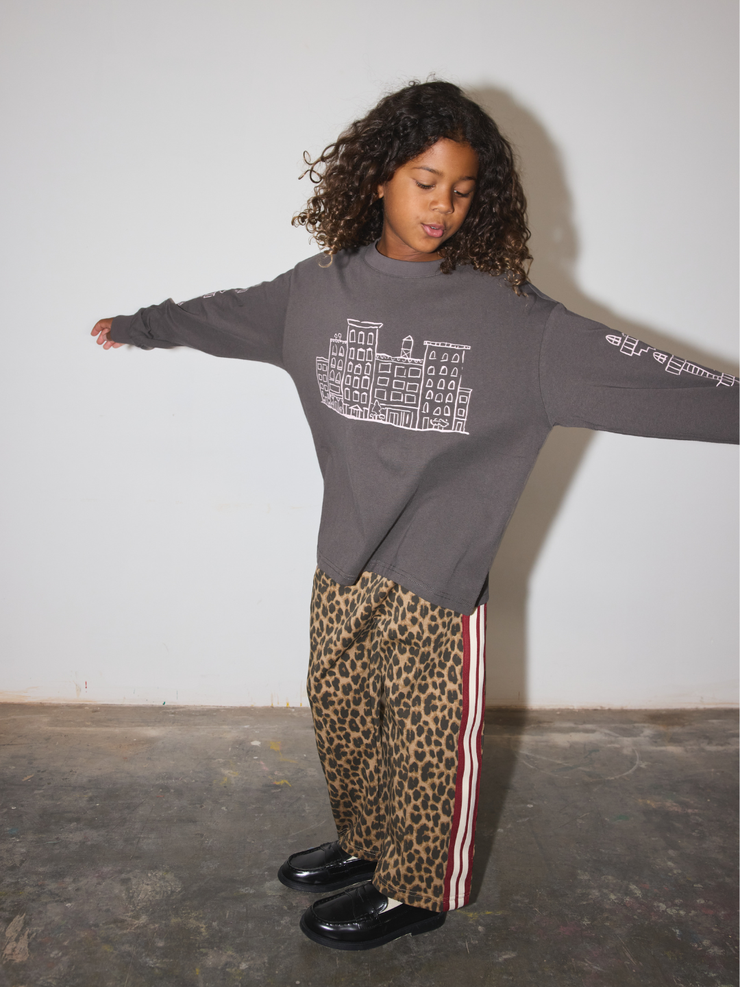 A child wearing a dark gray oversized sweatshirt with a white graphic print, paired with leopard print pants featuring red and white side stripes, standing against a plain white wall with arms outstreched.
