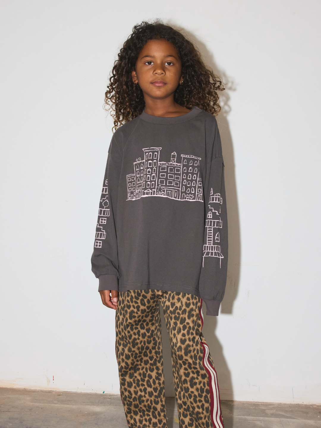 A young girl with curly dark hair stands against a plain off-white wall wearing a dark gray oversized sweatshirt with a white building graphic on the front and leopard-print pants with red and white side stripes.