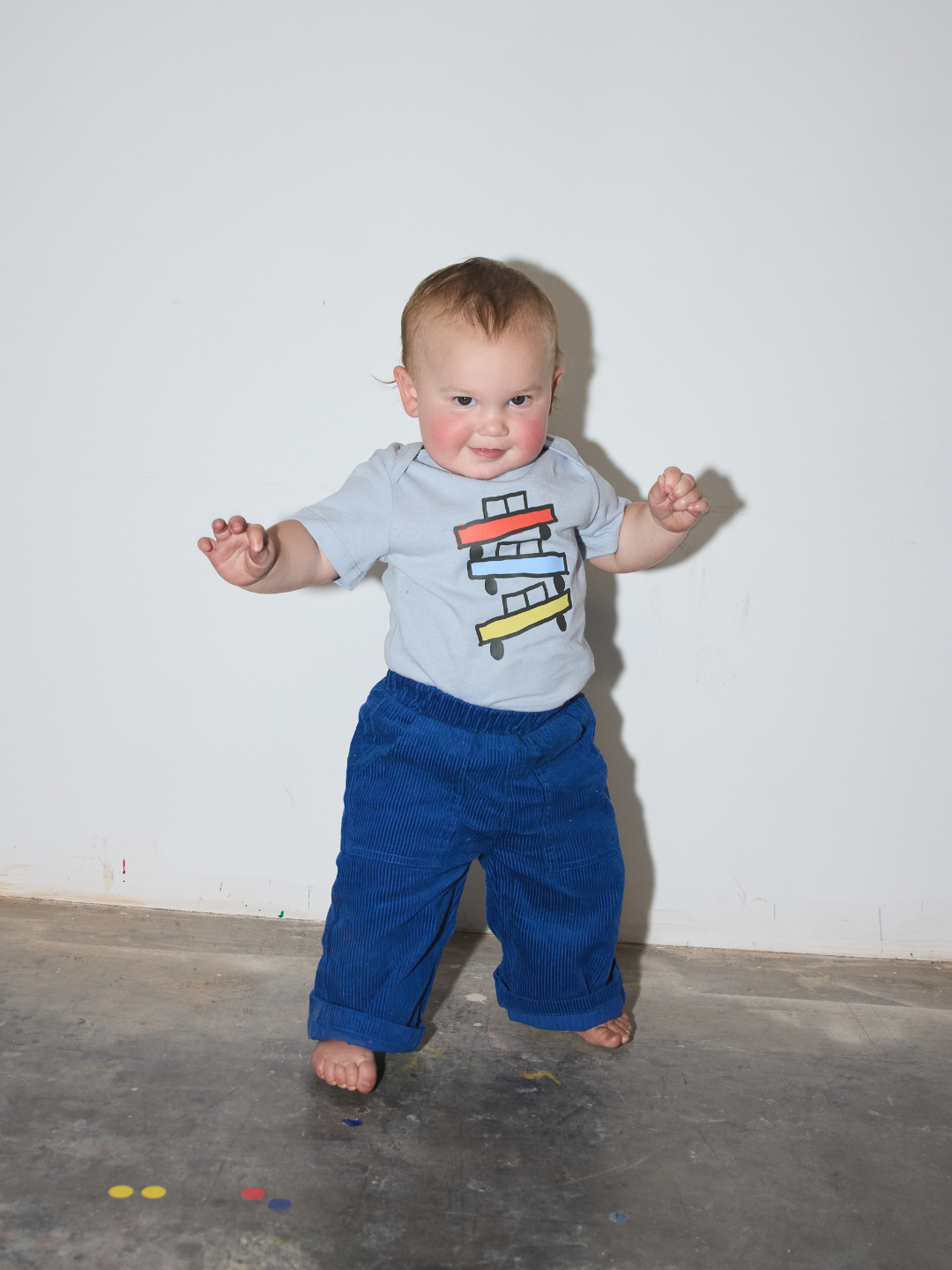 Blue | A baby standing barefoot on a concrete floor against a plain white wall, wearing a light gray t-shirt with a colorful airplane graphic and blue corduroy pants.
