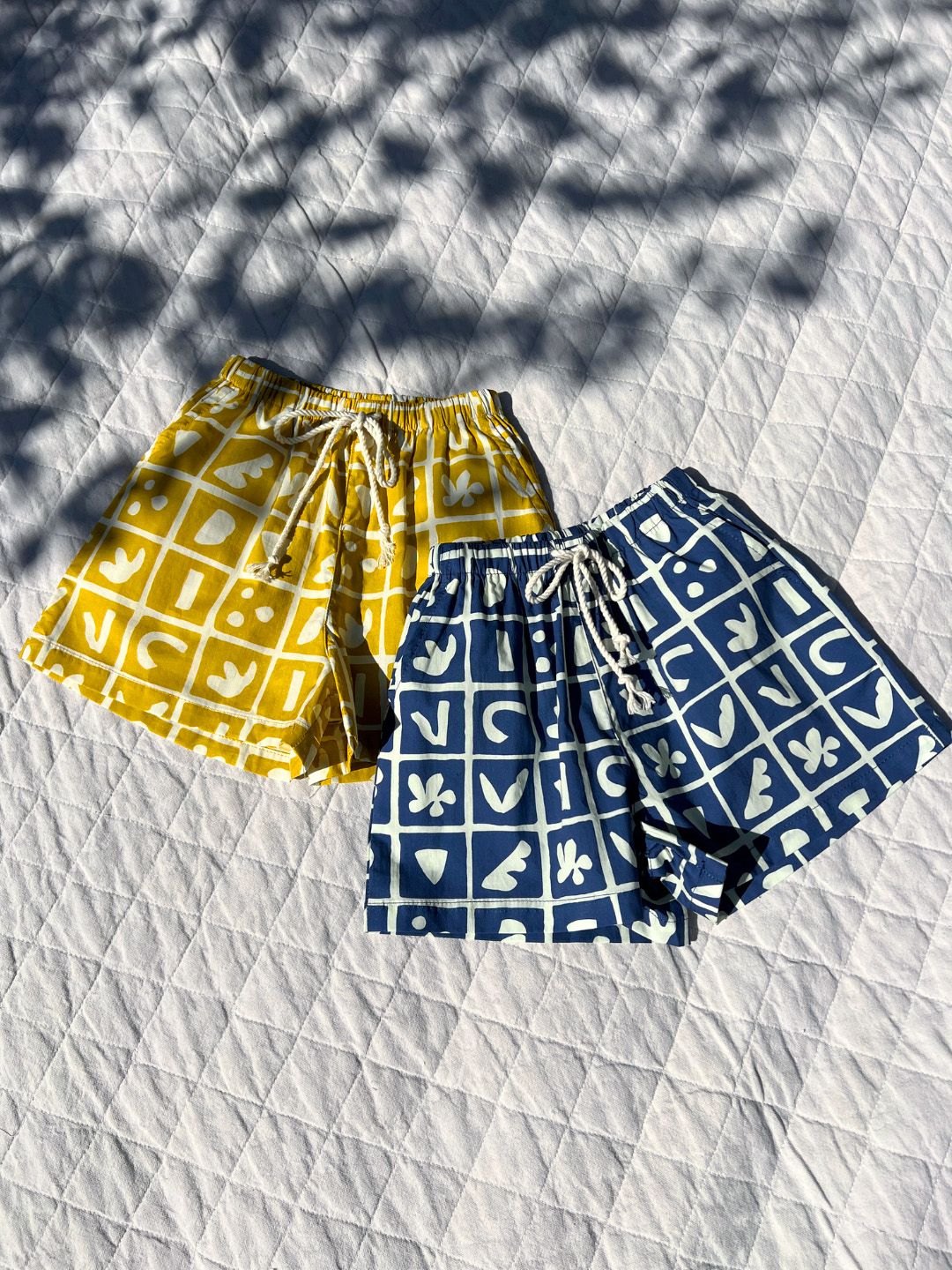 Blue | Two pairs of kids' shorts laid on a quilt; one in mustard yellow, one in deep blue, both overlaid with a grid of different shapes in white