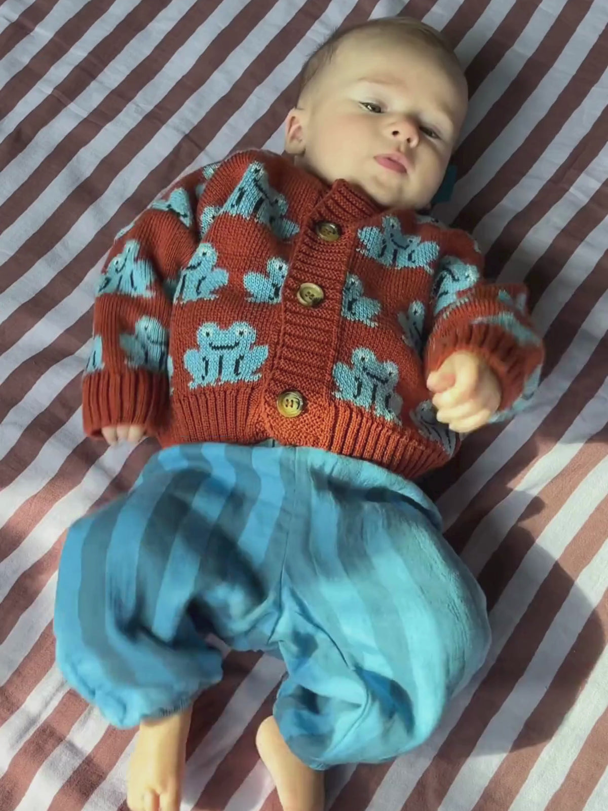 Video of a kicking baby wearing a red cardigan with a blue frogs motif, with blue striped pants, lying on a purple and burgundy striped quilt.