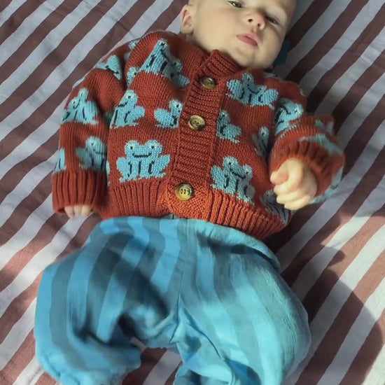 Blue Stripe | Video of a wiggly baby wearing the blue stripe organic cotton muslin pants with a red cardigan with blue frogs, lying on a purple striped quilt.
