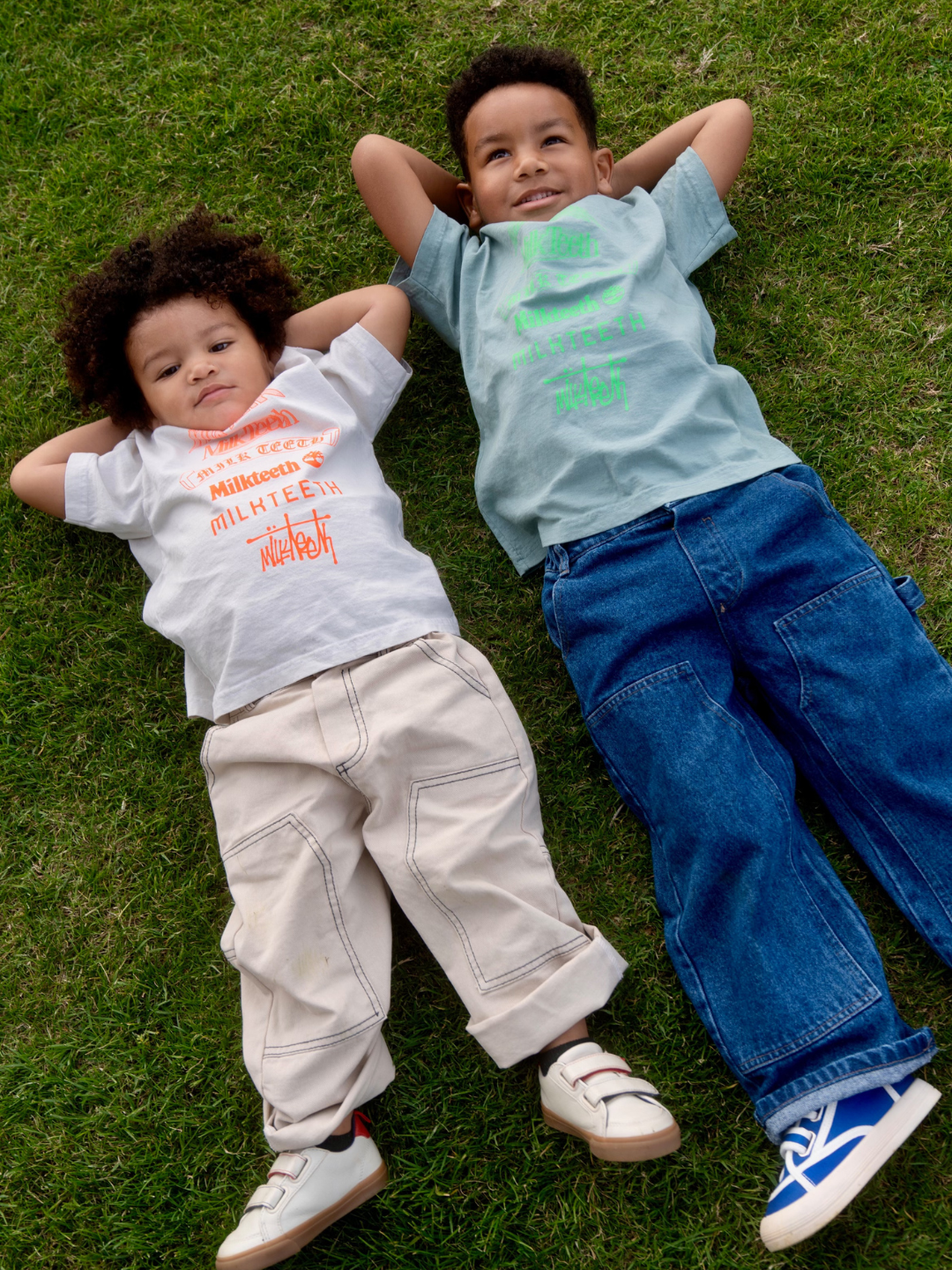 Green | Two children wearing a light green t-shirt with Milk Teeth spelled out in various fonts in neon green ink, and the same in beige. They wear jeans and and are relaxing on a lawn.