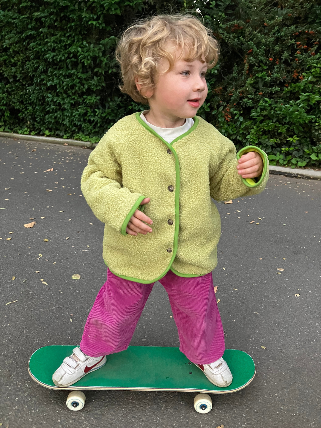 Child standing on a skateboard wearing magenta corduroy pants and a green fleece jacket, with a green hedge behind him, on a grey pavement.