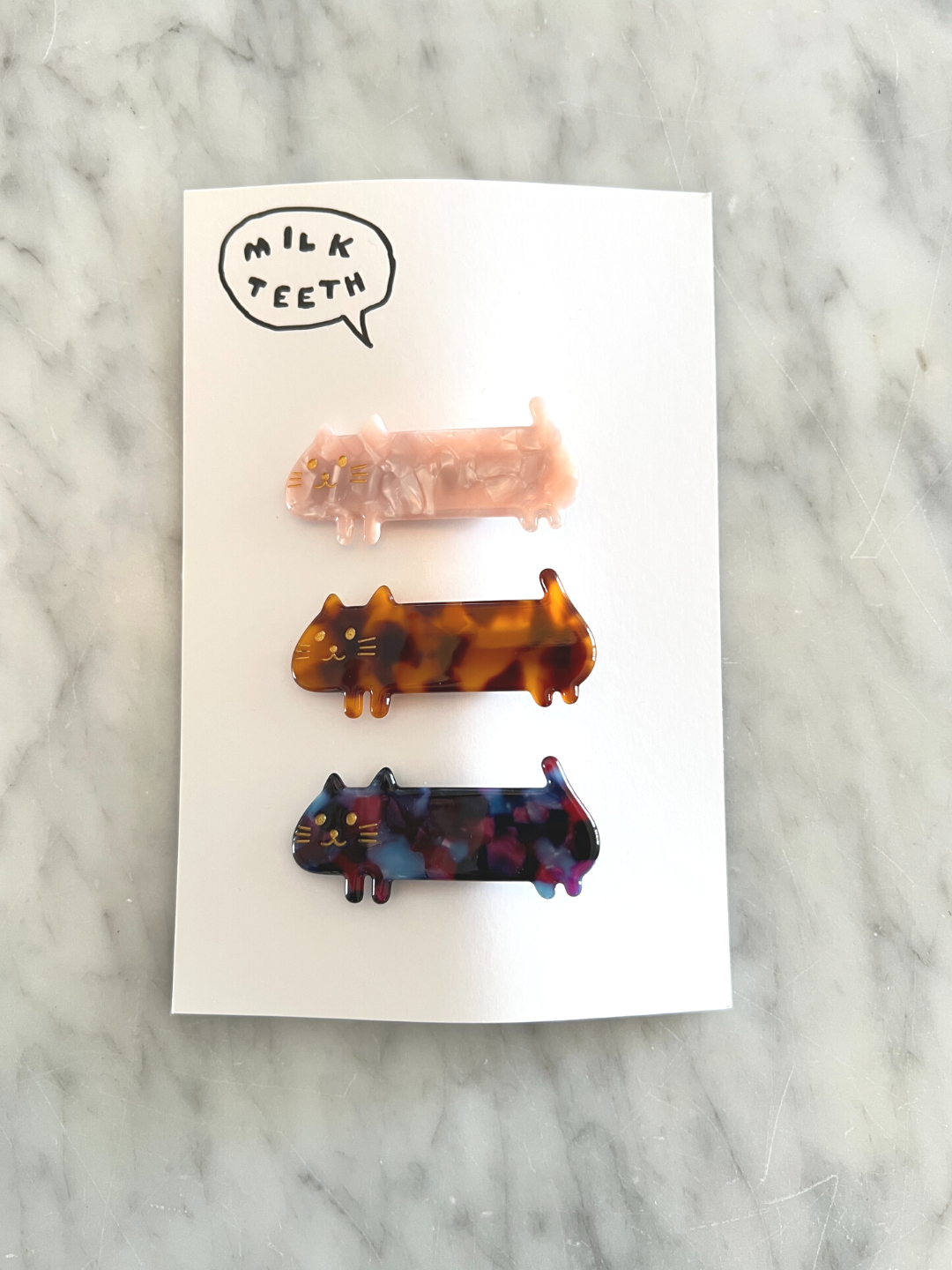 A Milk Teeth presentation card with three kids' barrettes in the shape of elongated cats: pink, ginger and purple