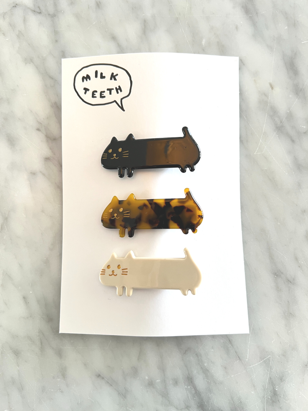 A Milk Teeth presentation card containing three kids' barrettes in the shape of elongated cats; one black, one tortoiseshell, one white