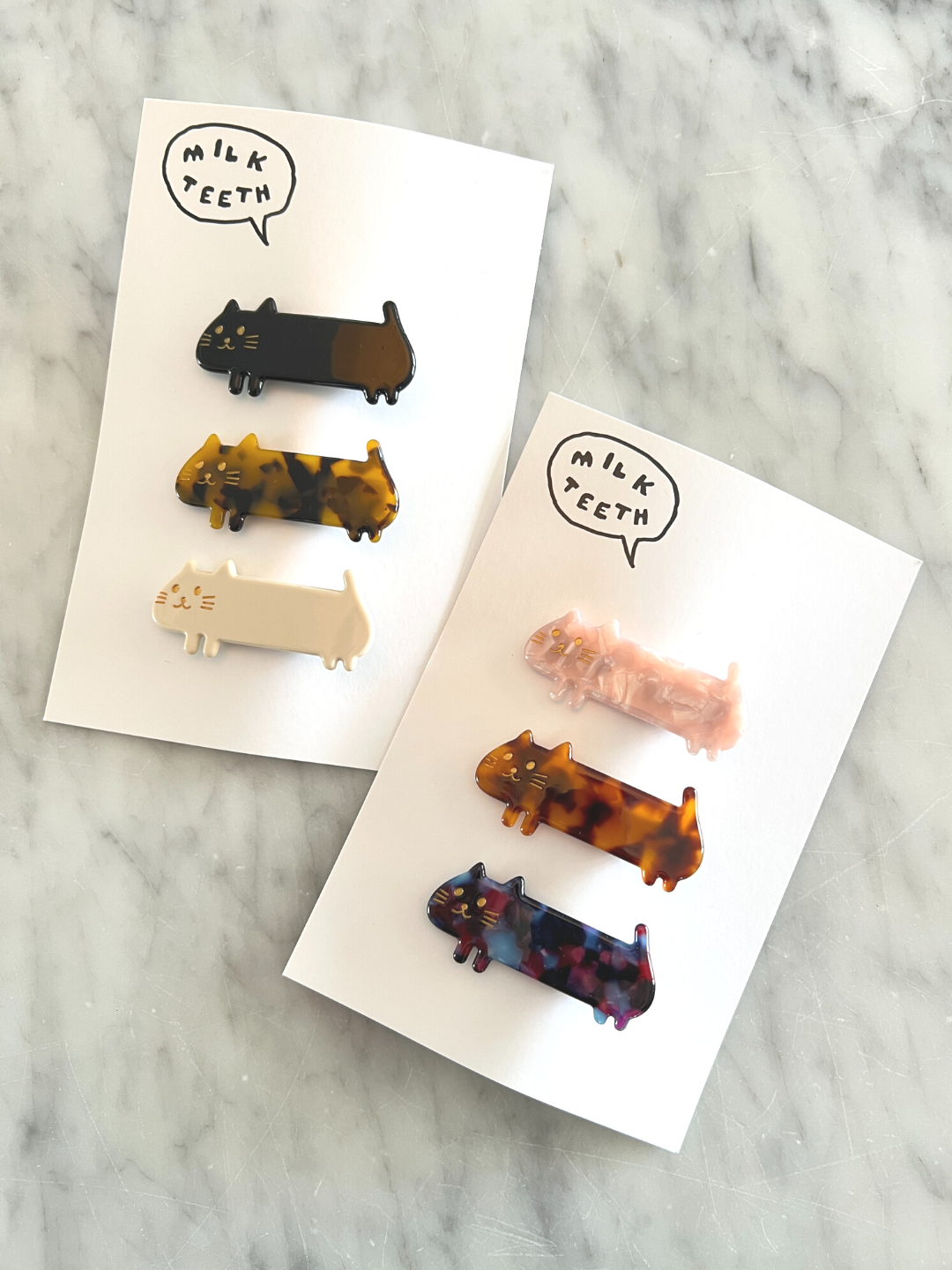 Two Milk Teeth presentation cards, each with three kids' barrettes in the shape of elongated cats; one set of black, tortoiseshell and white; one set of pink, ginger and purple