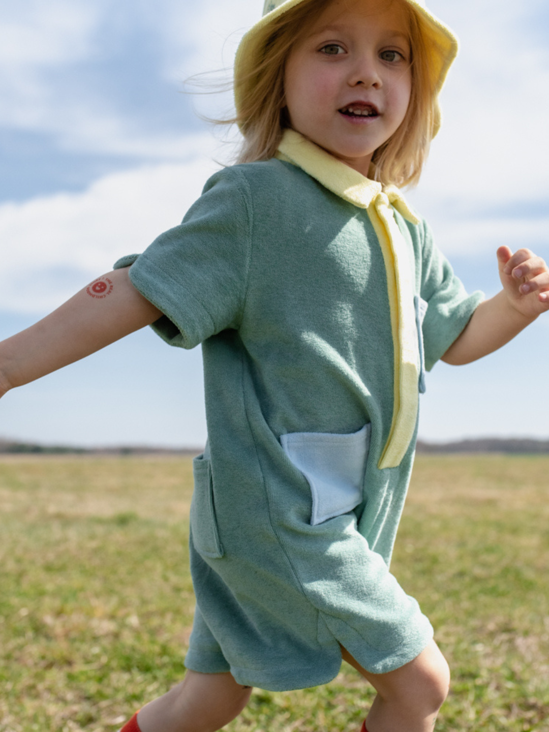 A child running in a pale green kids' jumpsuit with yellow collar and placket, blue pockets
