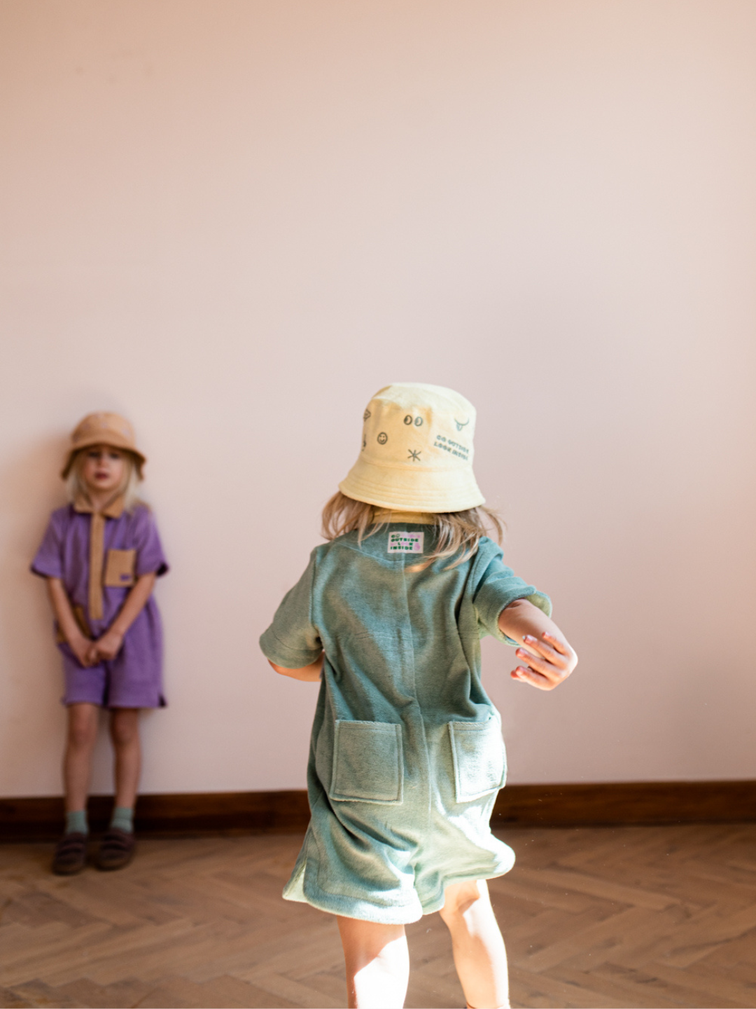 Two children in kids' jumpsuits: one in pale plum with sand collar and placket; one showing a rear view of pale green with yellow collar and two back pockets