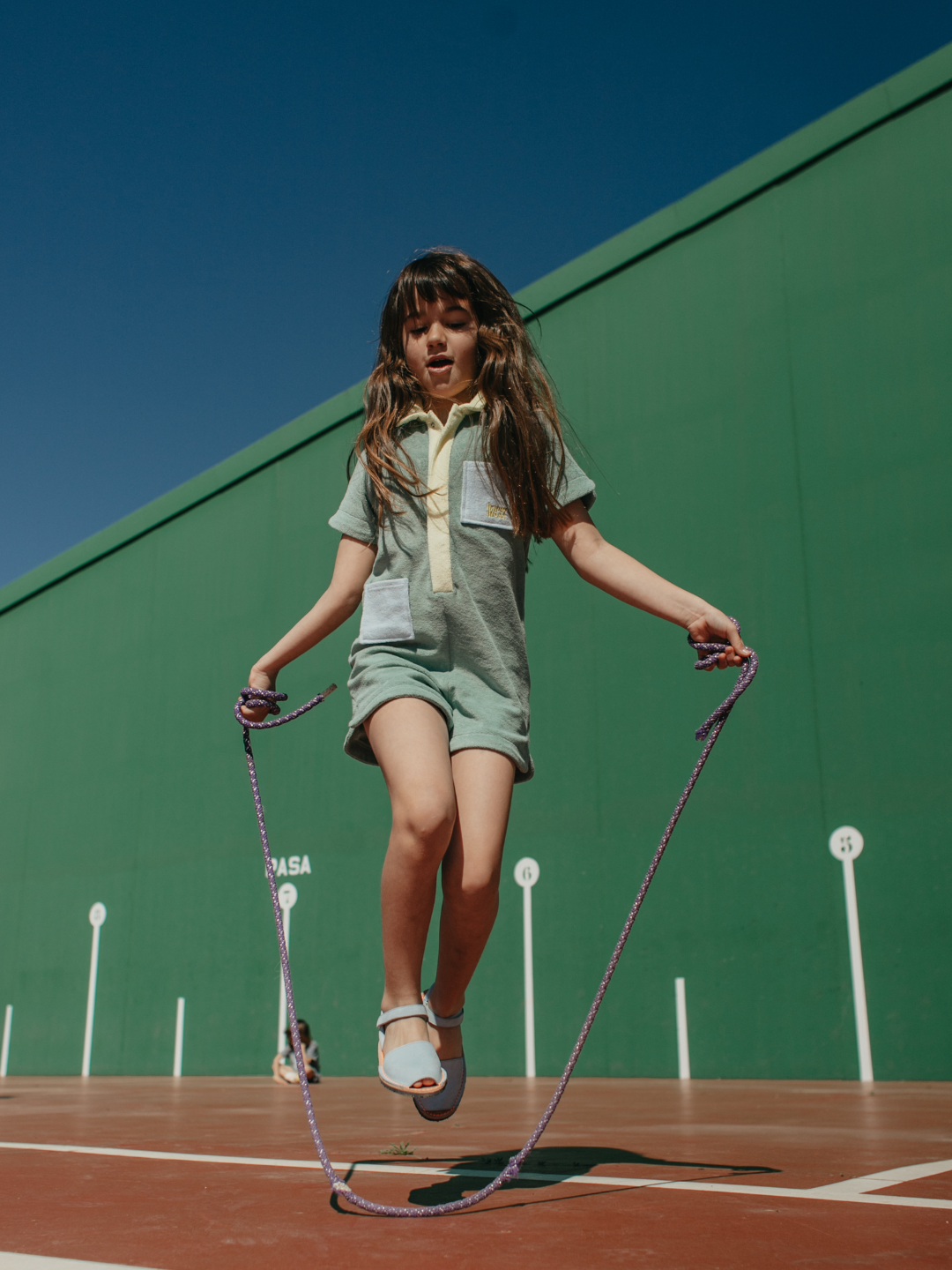 Cactus | A child skipping rope in a pale green kids' jumpsuit with yellow collar and placket and two blue pockets