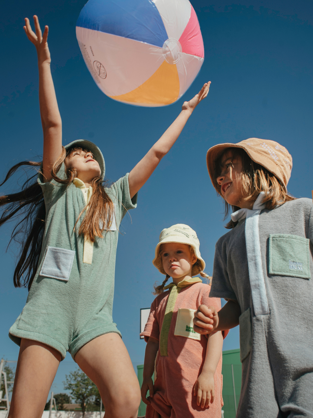 Cactus | Three children playing with a beachball wearing kids' jumpsuits in three different colors: one green with yellow collar and placket and blue pockets; one peach with green collar and placket, and yellow pockets; one pale gray with blue collar and placket, and green pockets