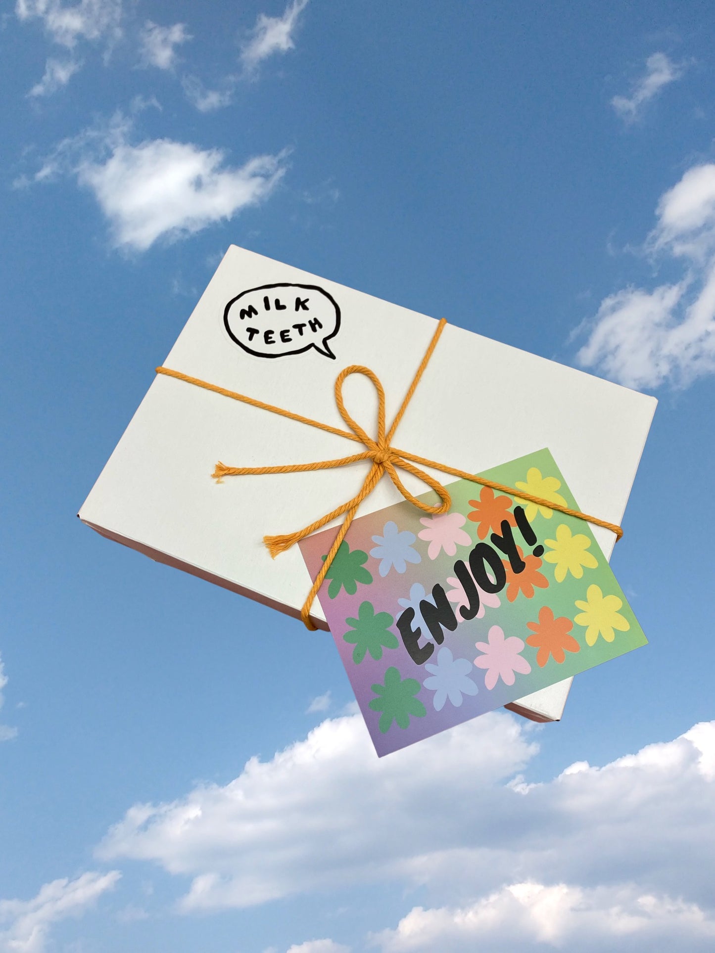 A gift box with yellow twine and Milk Teeth logo shown against a blue sky, with multicolored floral card