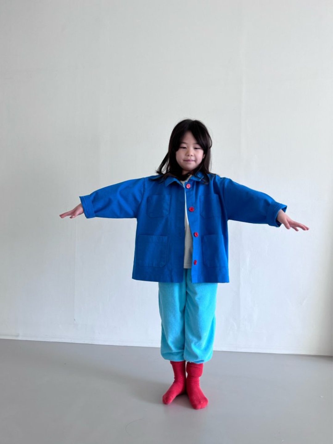A child wearing a kids' jacket in sky blue, with four pockets and four orange buttons, over turquoise pants