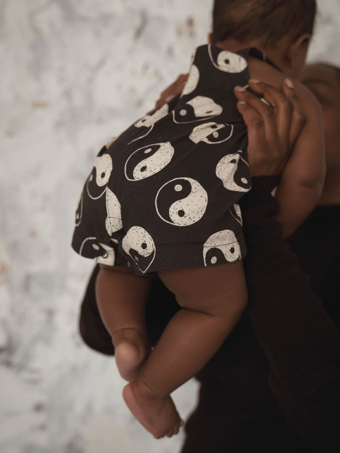 A back view of black overalls with a yin and yang pattern all over on a child being picked up by their parent.