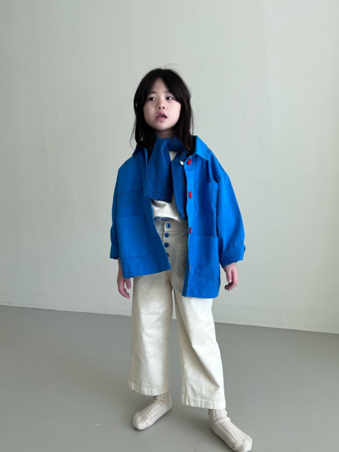 A child wearing a kids' jacket in sky blue, with four pockets and four orange buttons, over cream pants