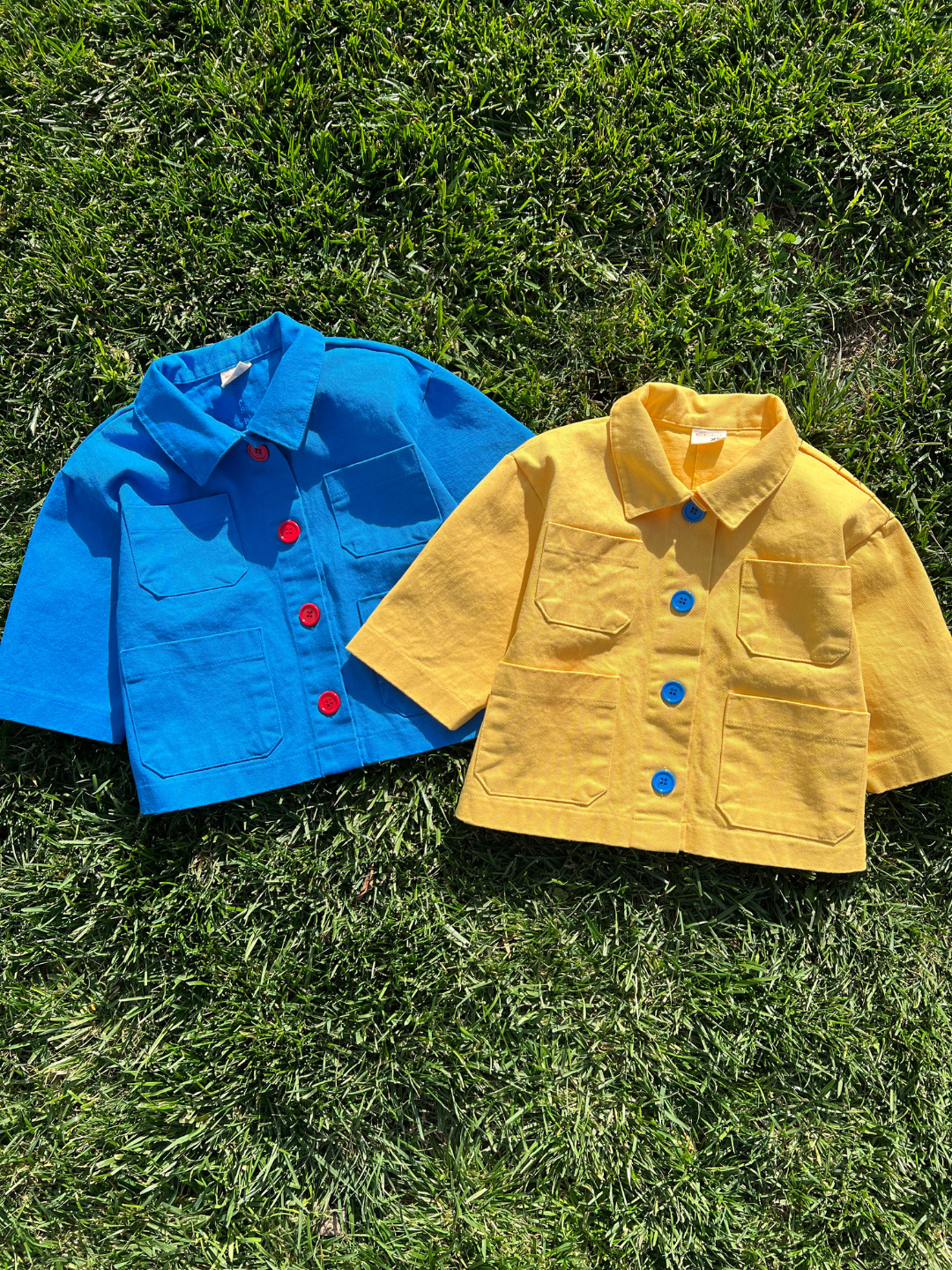 Yellow | Two kids' jackets laid on grass; one in sky blue with four pockets and four orange buttons, one in yellow with four pockets and four blue buttons