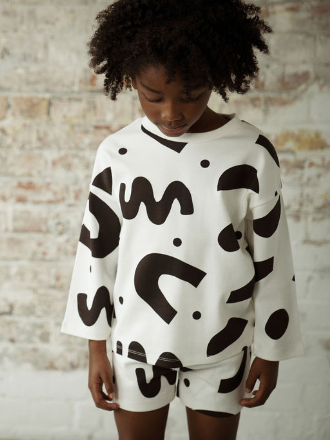 A child wearing a kids' tee long-sleeved shirt in a pattern of black squiggles and dots on a white background, with matching shorts