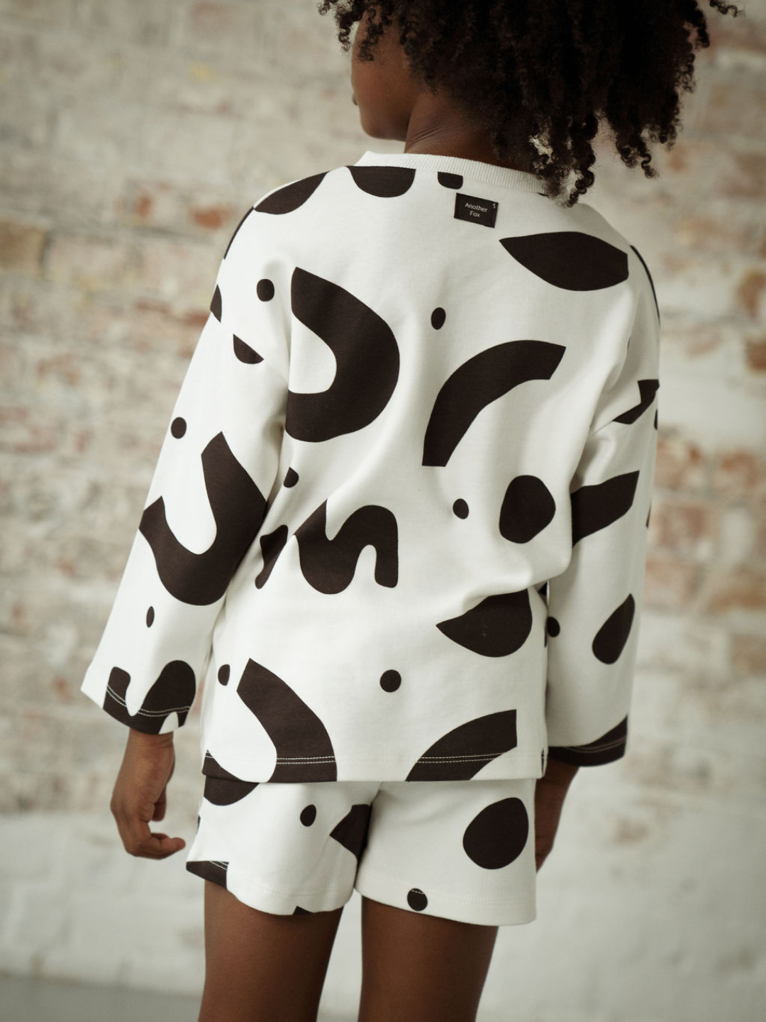 A child wearing a kids' long-sleeved tee shirt in a pattern of black squiggles and dots on a white background, with matching short, back view