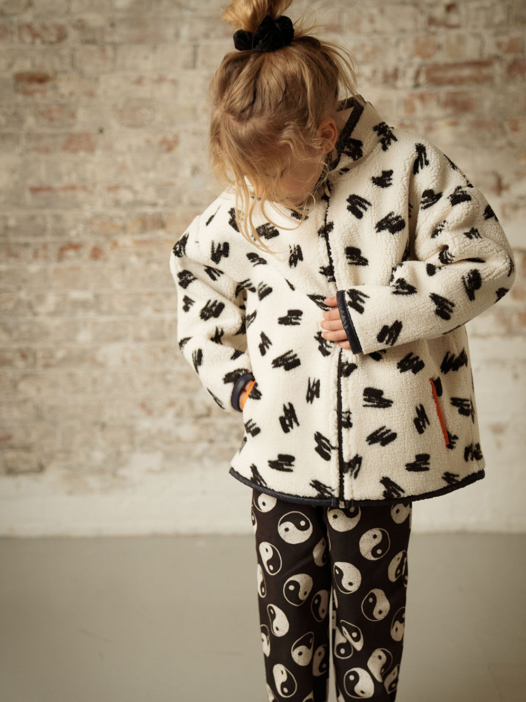A child wearing a kids' jacket in a pattern of black scribbles on a cream background, with black bindingon cuffs and hem, orange binding on pockets